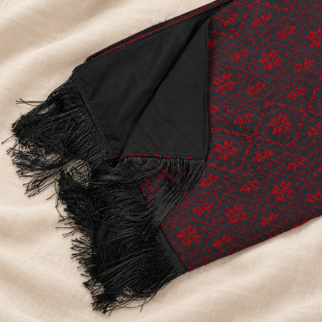 A close-up of a black scarf with red embroidery and black fringe, laid diagonally on a beige fabric background.