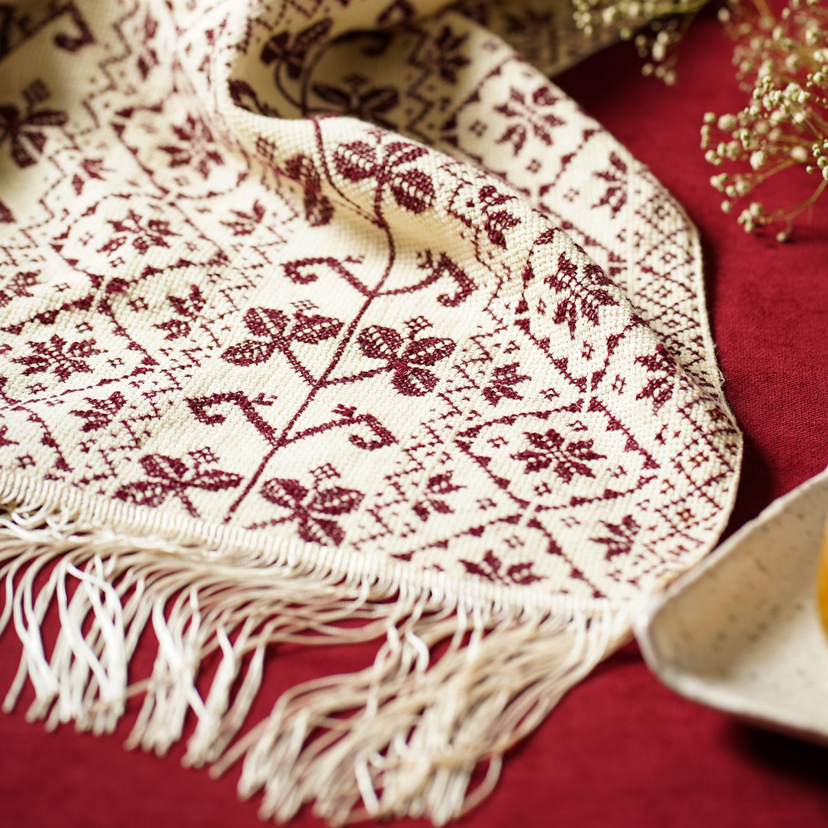 A cream-colored embroidered scarf with a dark red floral pattern and fringed edges, draped elegantly on a deep red fabric background with dried flowers and a ceramic dish as props.