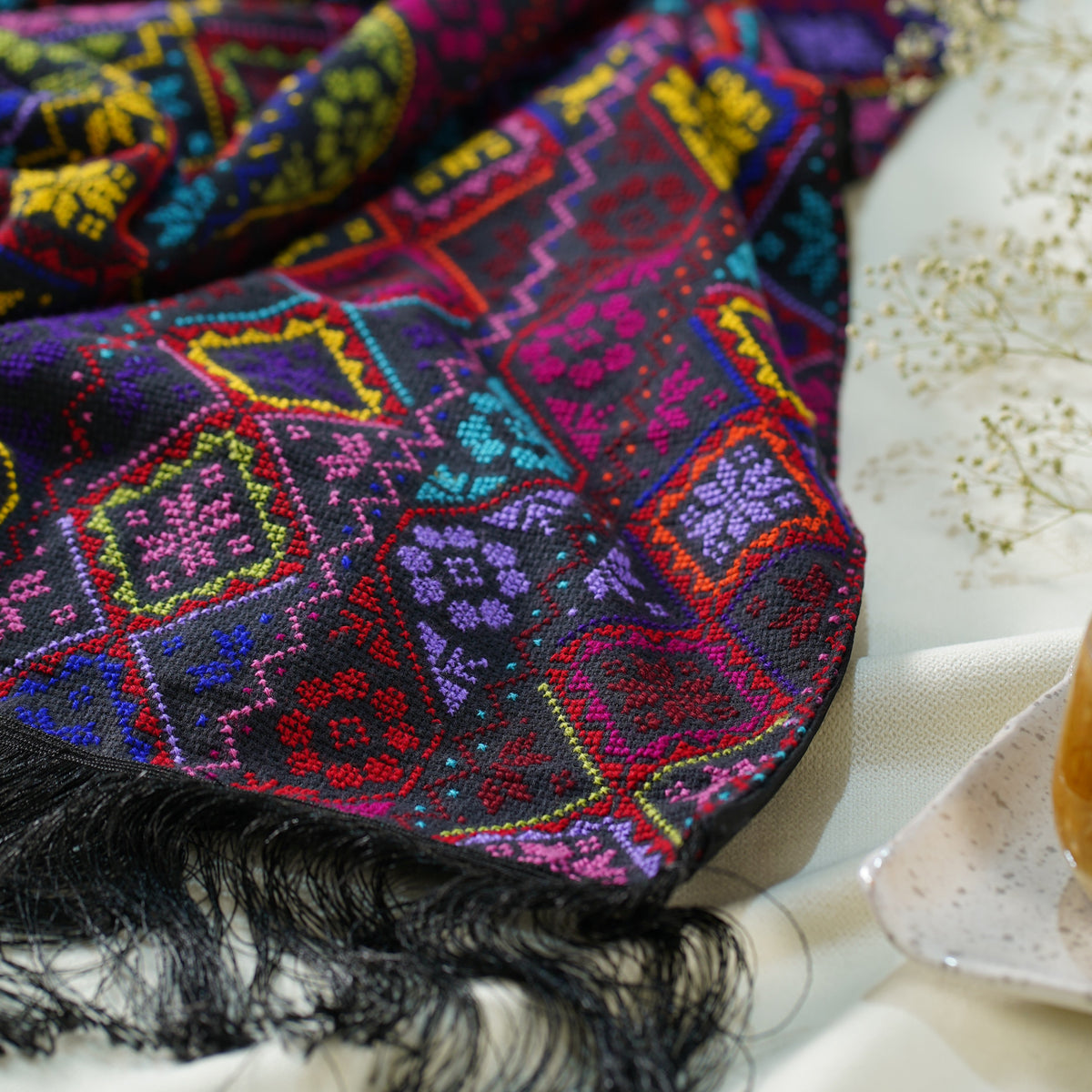 A close-up of a vibrant, multicolored embroidered shawl with black fringe, draped elegantly on a soft white fabric surface next to delicate dried flowers.