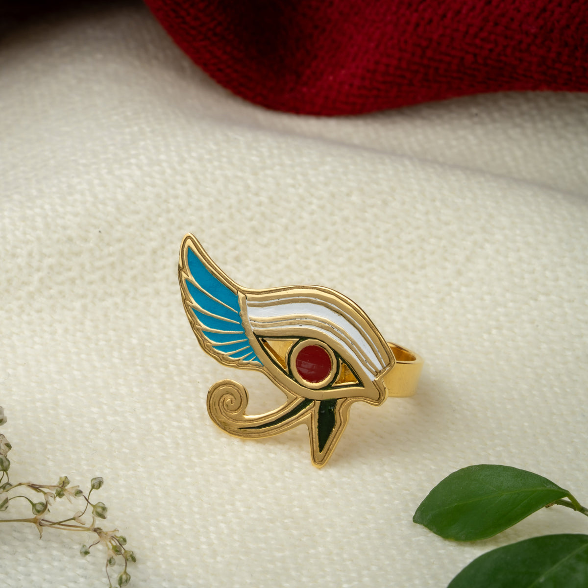 An Ancient Egyptian Eye of Horus ring with gold-plated metal, turquoise, red and white enamel, , displayed on a textured cream fabric with dried flowers and a red cloth in the background.