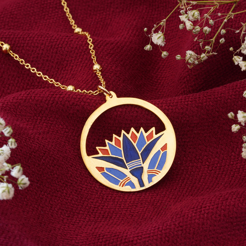 A gold circular pendant necklace featuring a stylized lotus flower in blue, red, and light blue enamel, displayed on a textured dark red fabric with white baby's breath flowers.