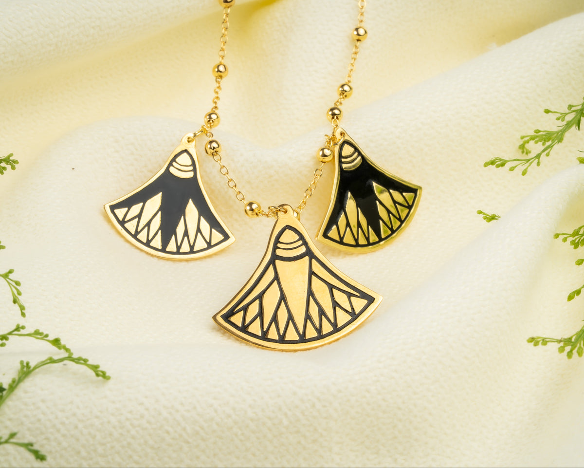 A gold chain necklace featuring three ancient Egyptian lotus pendant charms with black and gold patterns, displayed on a beige fabric and green botanic accent