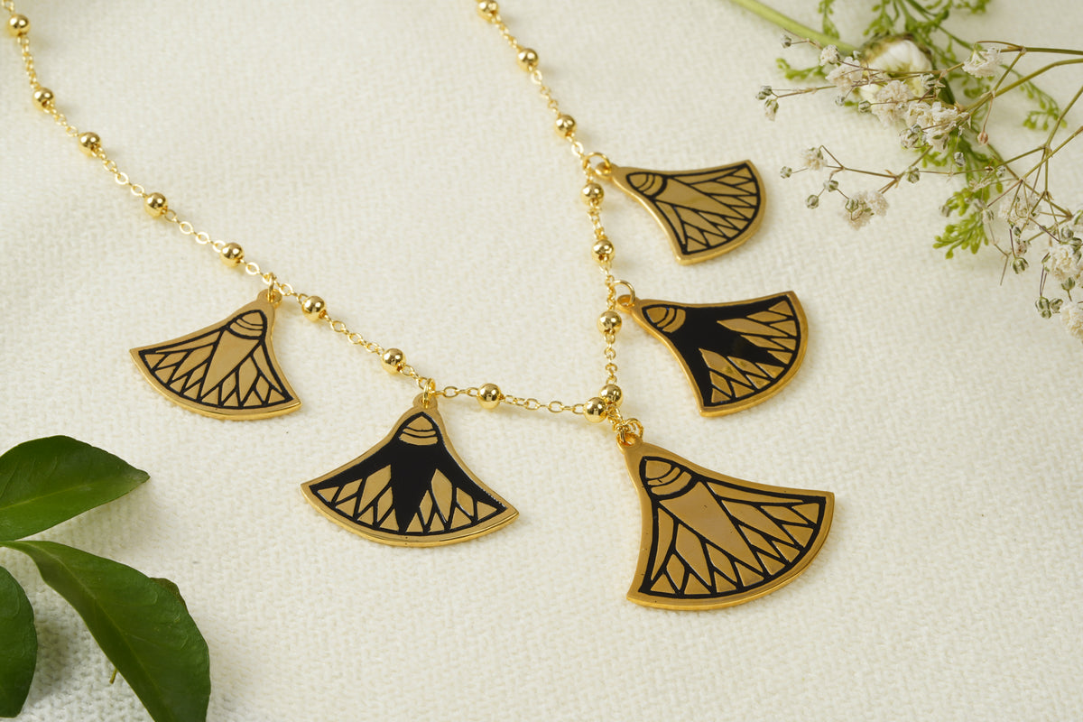 Gold statement necklace with five ancient egyptian lotus pendant charms featuring black and golden patterns, displayed on a textured white fabric with green leaves and baby's breath flowers.