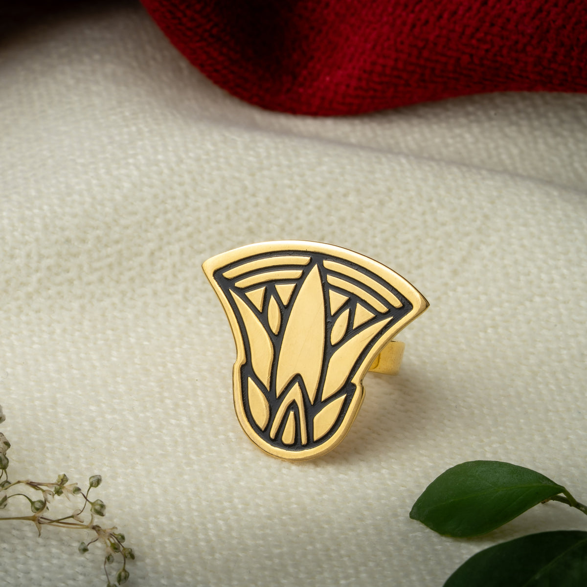 A gold plated lotus design inspired ring styled on a textured cream fabric with a maroon knit fabric, baby's breath flowers, and green leaves.
