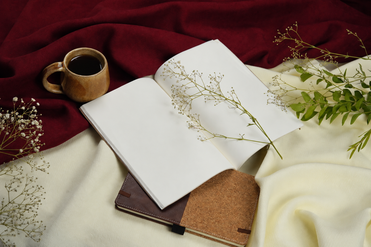An open blank notebook with white pages lies on draped cream and deep red fabrics, with a small wooden mug of black coffee, dried flowers, and greenery around it.