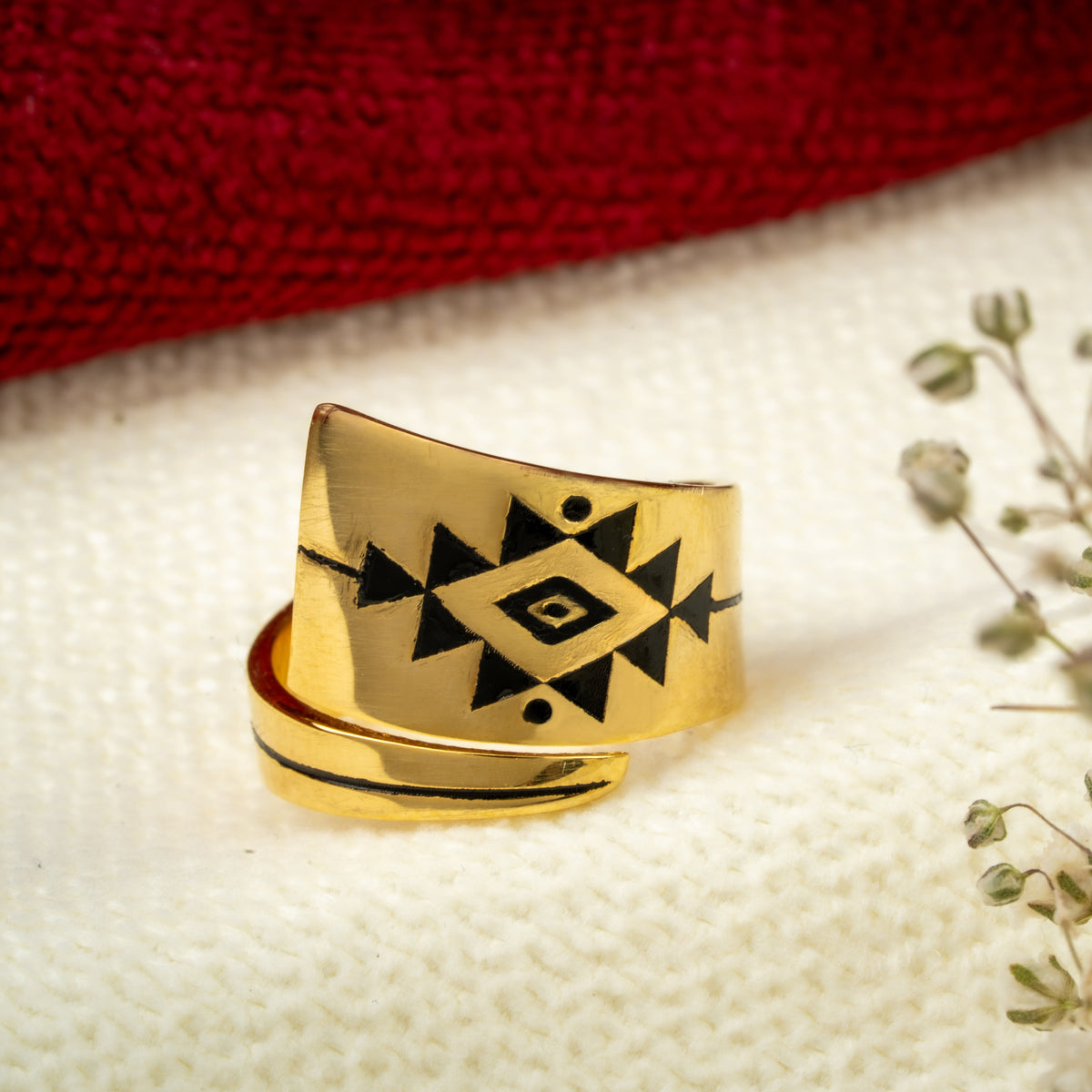 A gold wrap ring with a black geometric diamond pattern, displayed on a textured white surface next to small white flowers and a red fabric background.