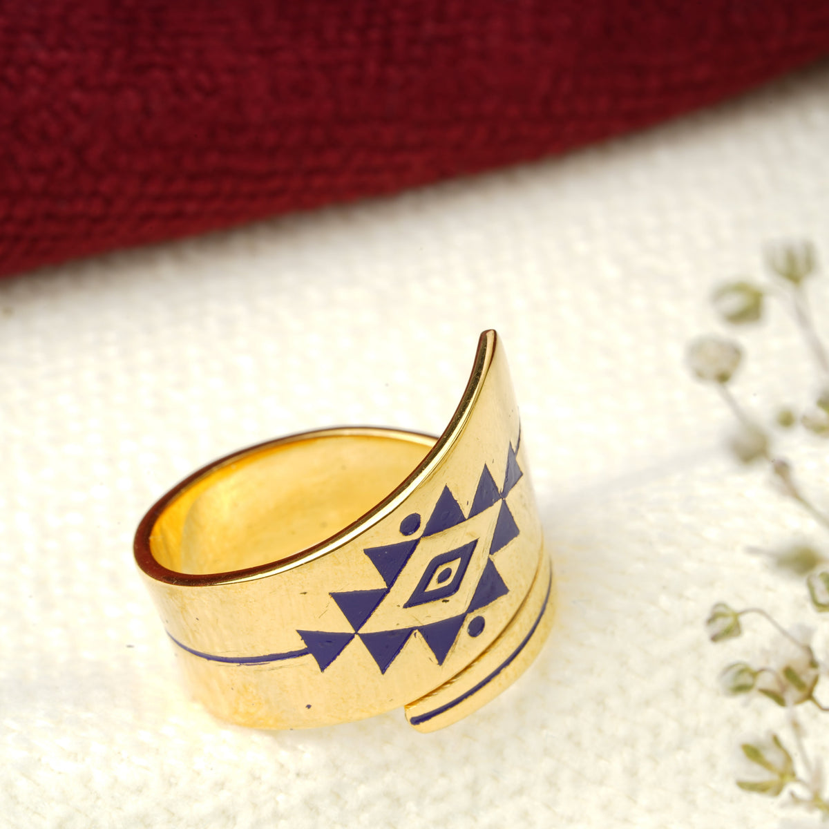A wrap band ring with a violet geometric diamond pattern, displayed on a textured white surface next to small white flowers and a dark red fabric background.