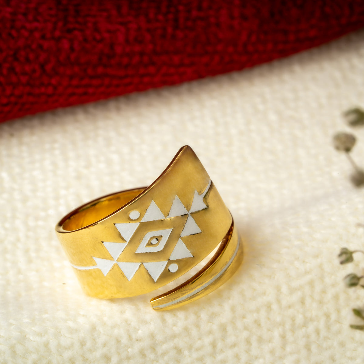 A gold wrap ring with a white geometric diamond pattern, resting on a textured cream fabric next to dried baby's breath flowers.