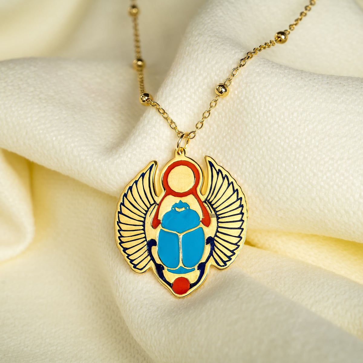 A gold-plated pendant necklace featuring an Egyptian scarab beetle design with a blue body, red accents, and navy wing details, resting on draped cream-colored fabric.