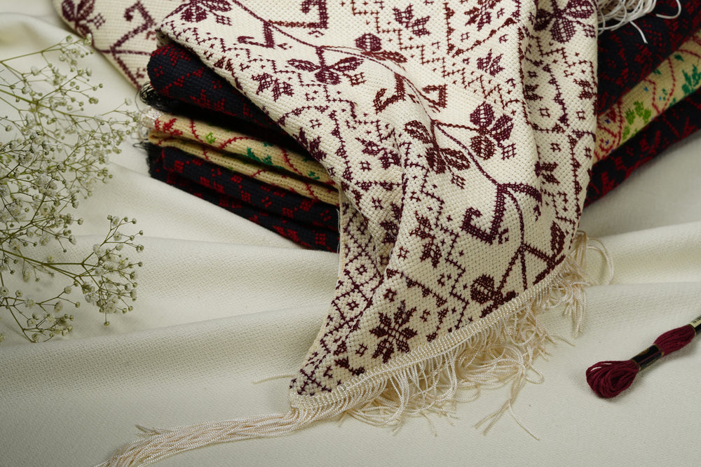 A close-up of a folded embroidered shawl with cream ground and dark burgundy floral and geometric embroidery, draped over other embroidered shawl on a light fabric surface. A small tassel is visible at the lower right, with delicate white flowers as a prop on the left.