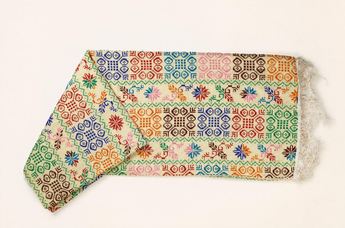 A folded multicolor patterned scarf with white fringe, displayed on a clean off-white background.