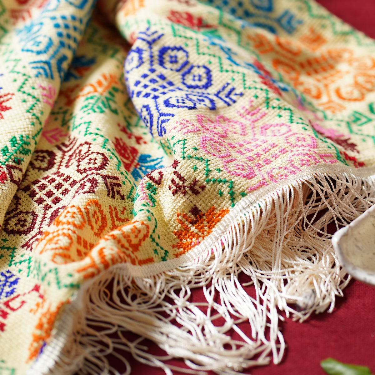 A close-up of a multicolored patterned scarf with cream fringe, draped on a deep red fabric background.
