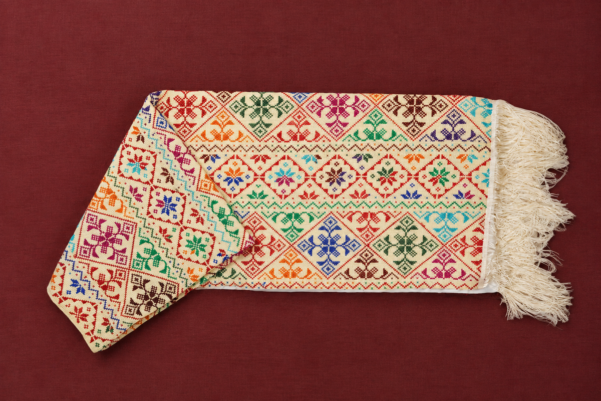 A shawl with cream fringed edges and a multicolored geometric and floral pattern, laid flat on a dark red background.