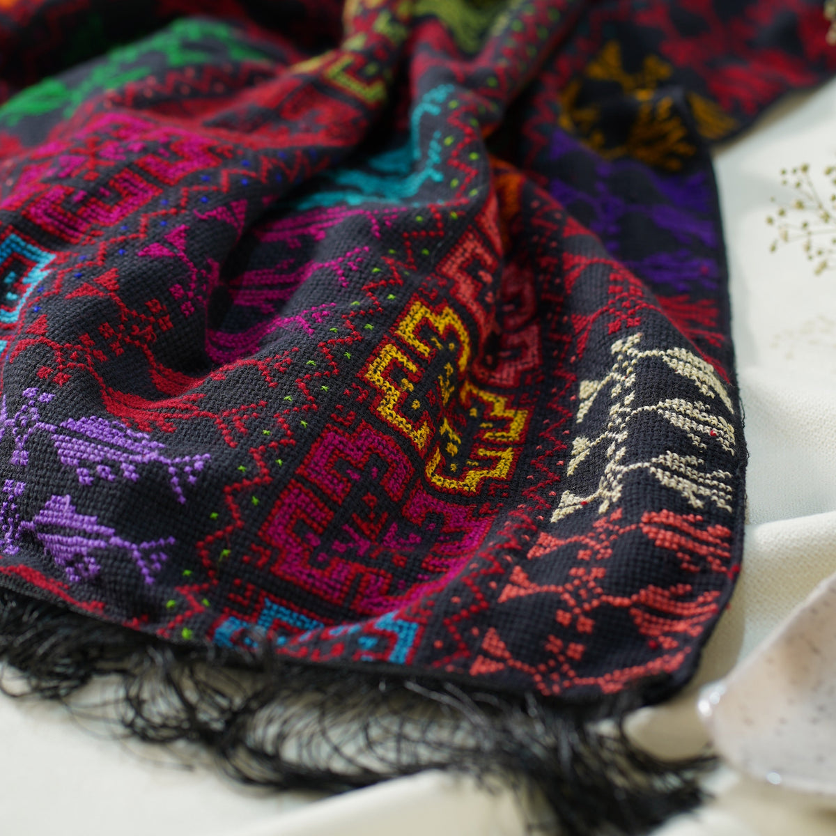 A close-up of a multicolored embroidered scarf with black fringe, featuring intricate geometric and floral patterns in red, purple, blue, green, and yellow on a dark background, styled on a white fabric surface.