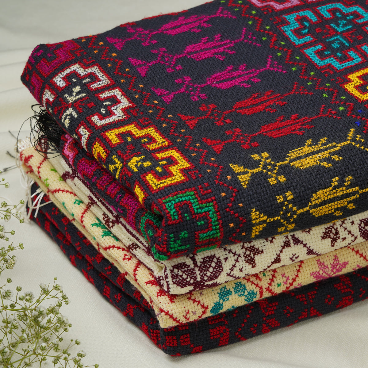 A stack of folded embroidered shawls with colorful geometric and floral patterns, styled with small white flowers and a pale peach rose on a light fabric background.