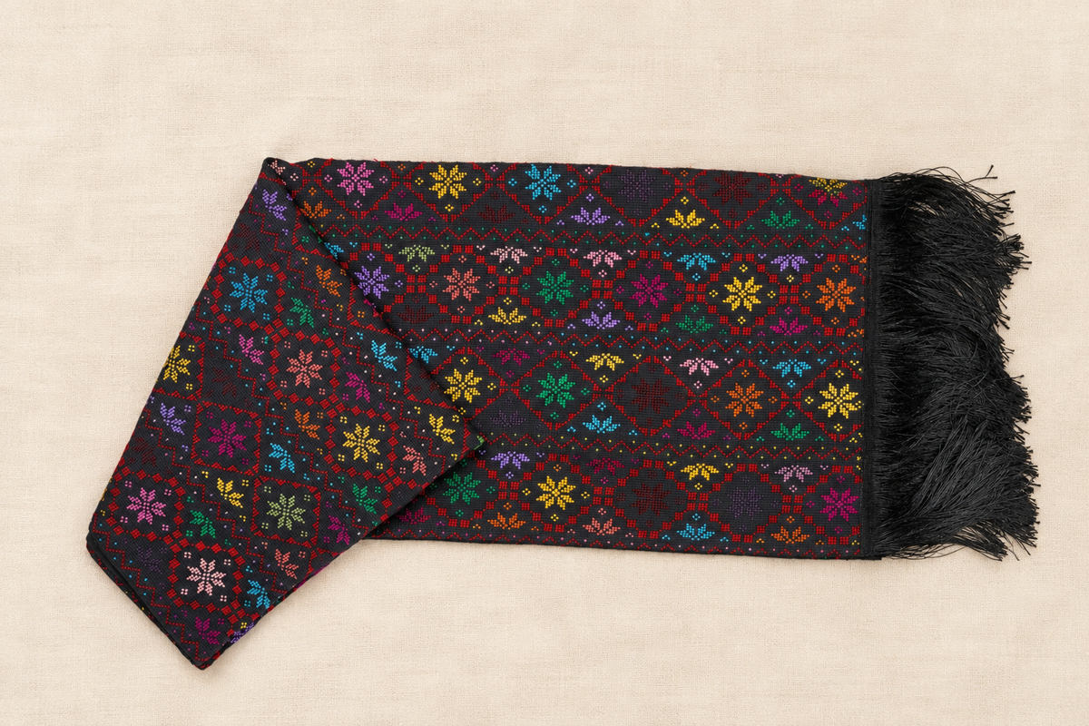 A folded black scarf with colorful geometric and floral embroidery, finished with black fringe, displayed on a light beige fabric background.