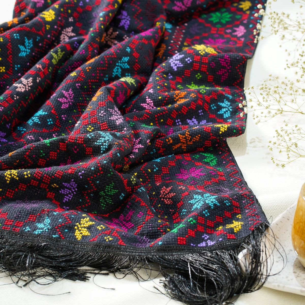 A hand embroidered shawl with intricate multicolor geometric and floral patterns, finished with black fringe  draped on a white surface.