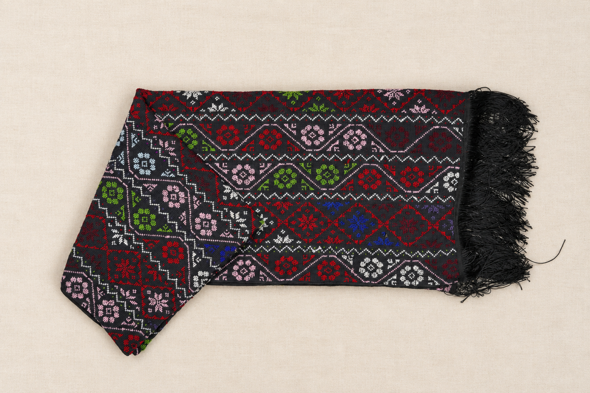 A folded black shawl  with colorful geometric and floral embroidery patterns in red, green, pink, blue, and white, finished with black fringe on one end, laid flat on a light beige background.