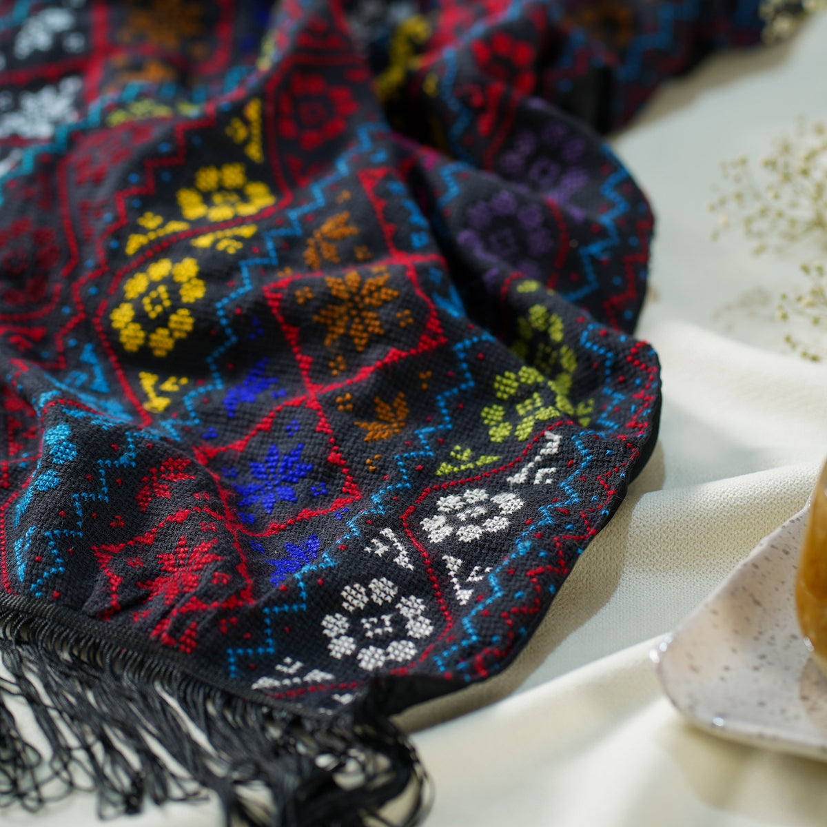 A close-up of a scarf with black fringe, featuring a vibrant multicolor geometric and floral embroidery in red, blue, yellow, and white on a dark background, styled on a draped white fabric surface.