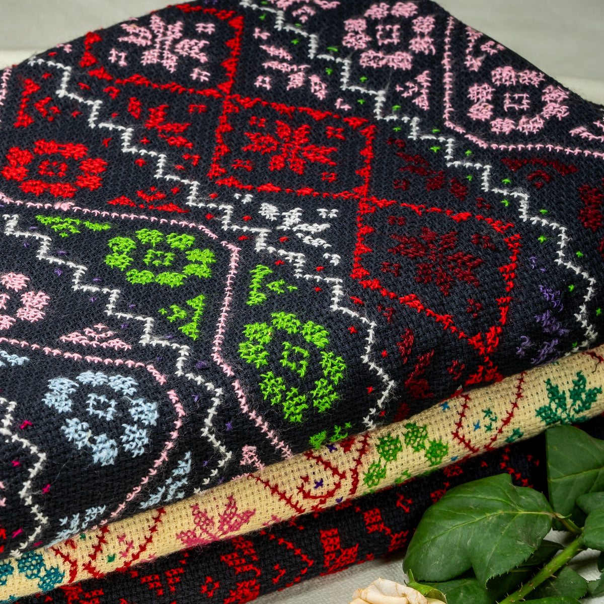 A stack of folded patterned shawls, with the top piece prominently showing a black base and colorful geometric and floral motifs in red, pink, green, blue, and white. The textiles are styled on a light fabric background with small green leaves in the corners.