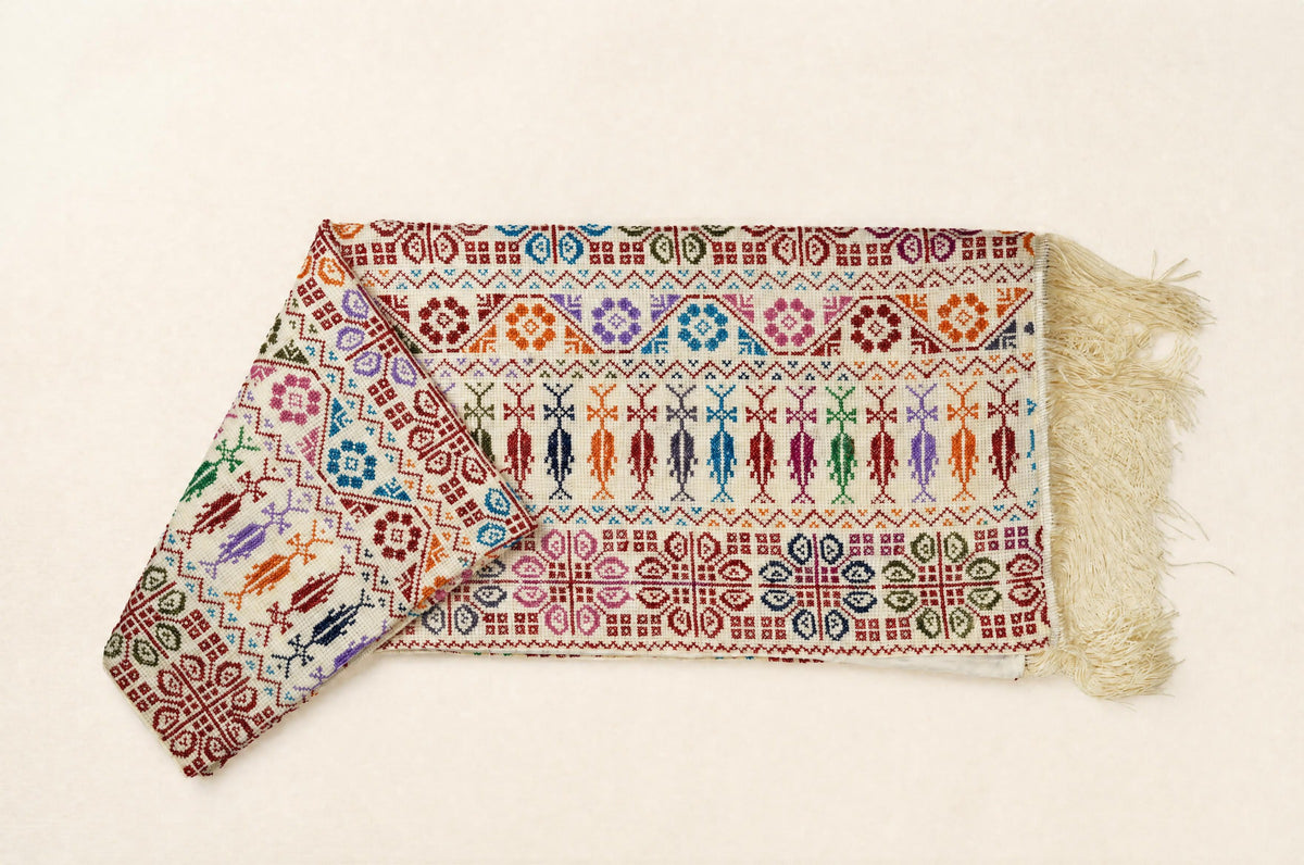 A folded embroidered scarf with cream-colored fringe, featuring intricate geometric and tribal-style embroidery in red, blue, green, orange, and purple on a light beige background.