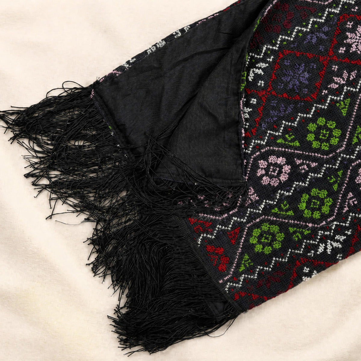 A close-up of a black embroidery scarf with fringe, featuring a colorful geometric and floral pattern in red, green, pink, and white on a cream fabric background.