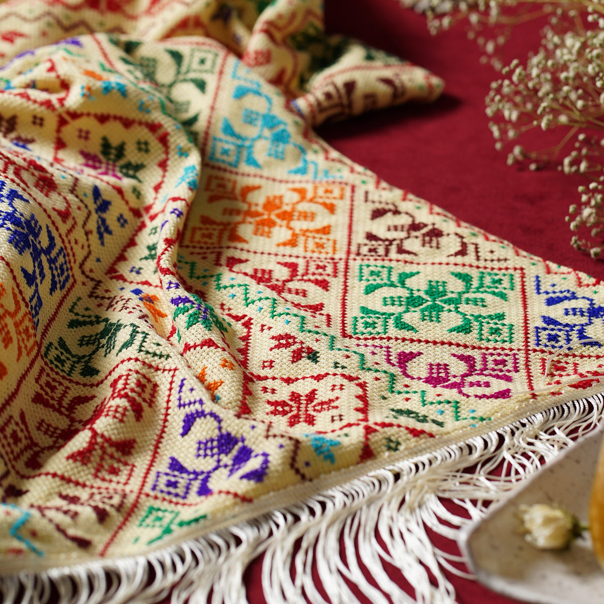 A close-up of a cream-colored embroided shawl with colorful geometric and floral patterns, draped on a deep red fabric background with white tassels along one edge.