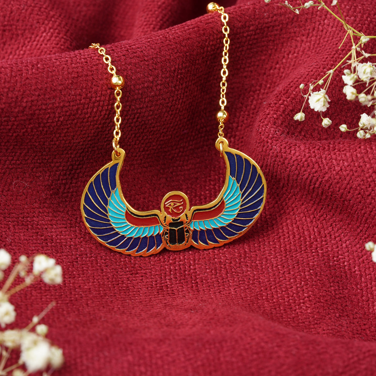 Gold-plated ancient egyptian themed necklace with a winged scarab beetle pendant in blue, teal, red and gold, displayed on a textured dark red fabric with baby's breath flowers.