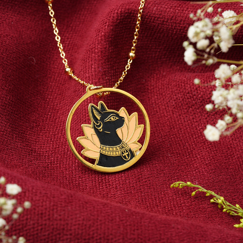 A gold pendant necklace featuring a Ancient Egyptian goddess Bastet profile with gold lotus petals and an Ankh symbol, displayed on a textured dark red fabric with white baby's breath flowers.