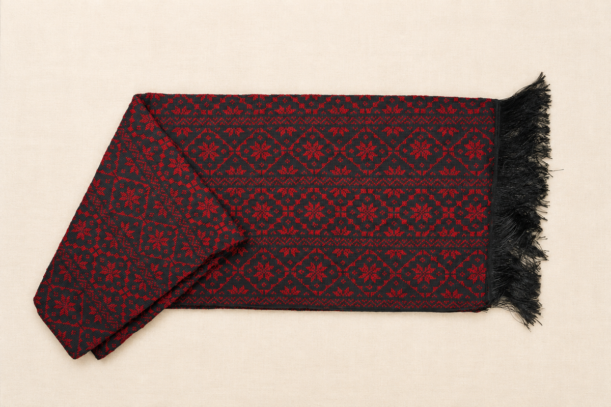 A folded black scarf with a red geometric and floral hand embroidered pattern, featuring black fringe , laid flat on a light beige background.
