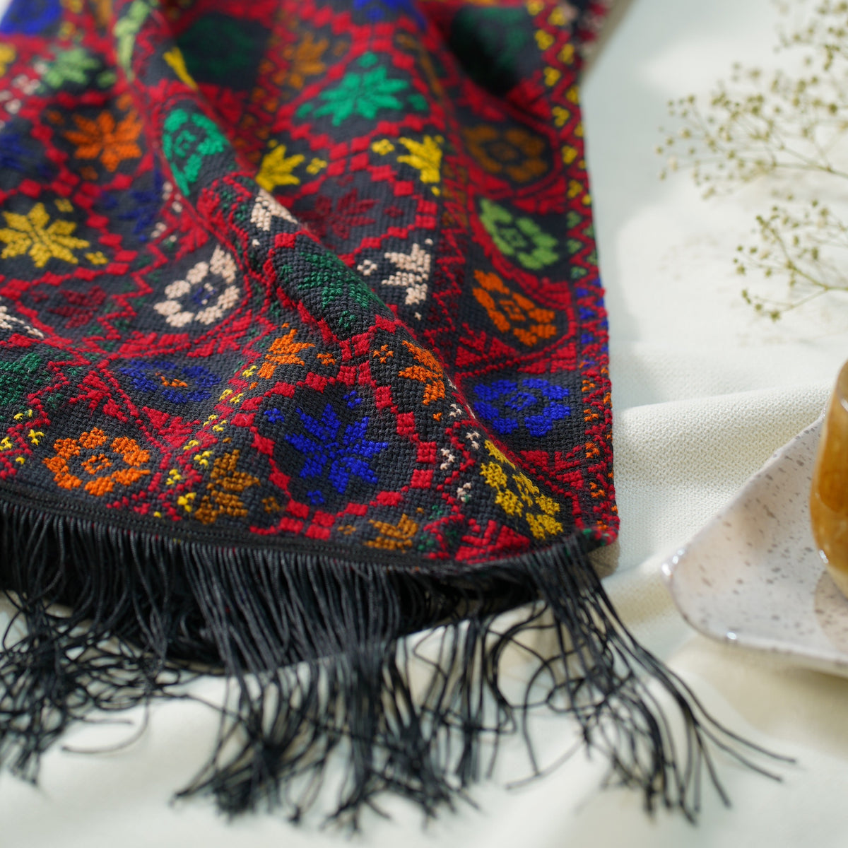 A close-up of hand embroidered shawl  with black fringe, featuring a vibrant multicolor geometric and floral pattern on a dark background, styled on a white textured surface.