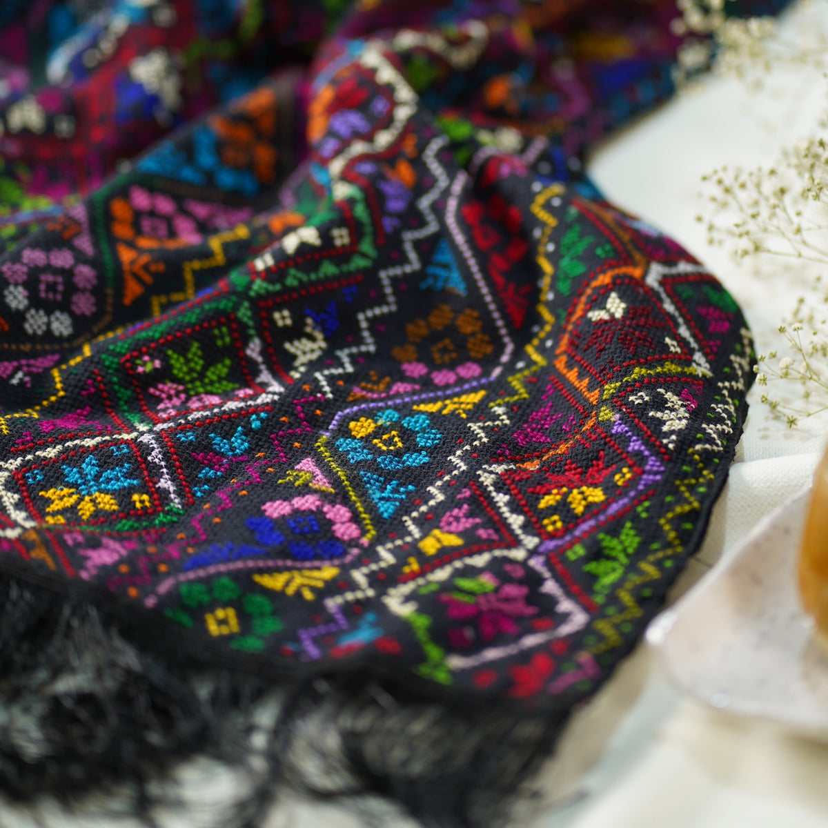A close-up of a dark, intricately hand embroidered scarf with black fringe, featuring vibrant multicolor geometric and floral patterns in purple, blue, green, yellow, and red, styled on a light surface with dried flowers.