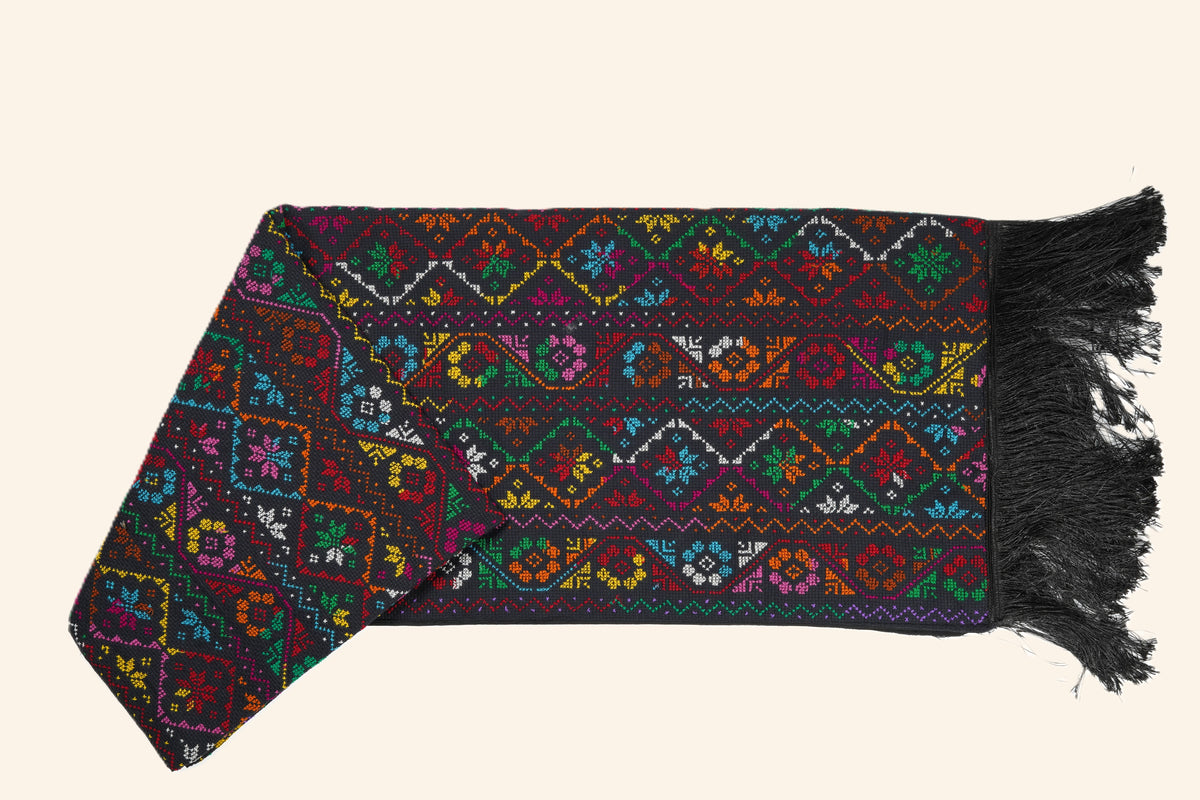 A hand embroidered shawl with intricate multicolor geometric and floral patterns, finished with black fringe , displayed on a solid light beige background.