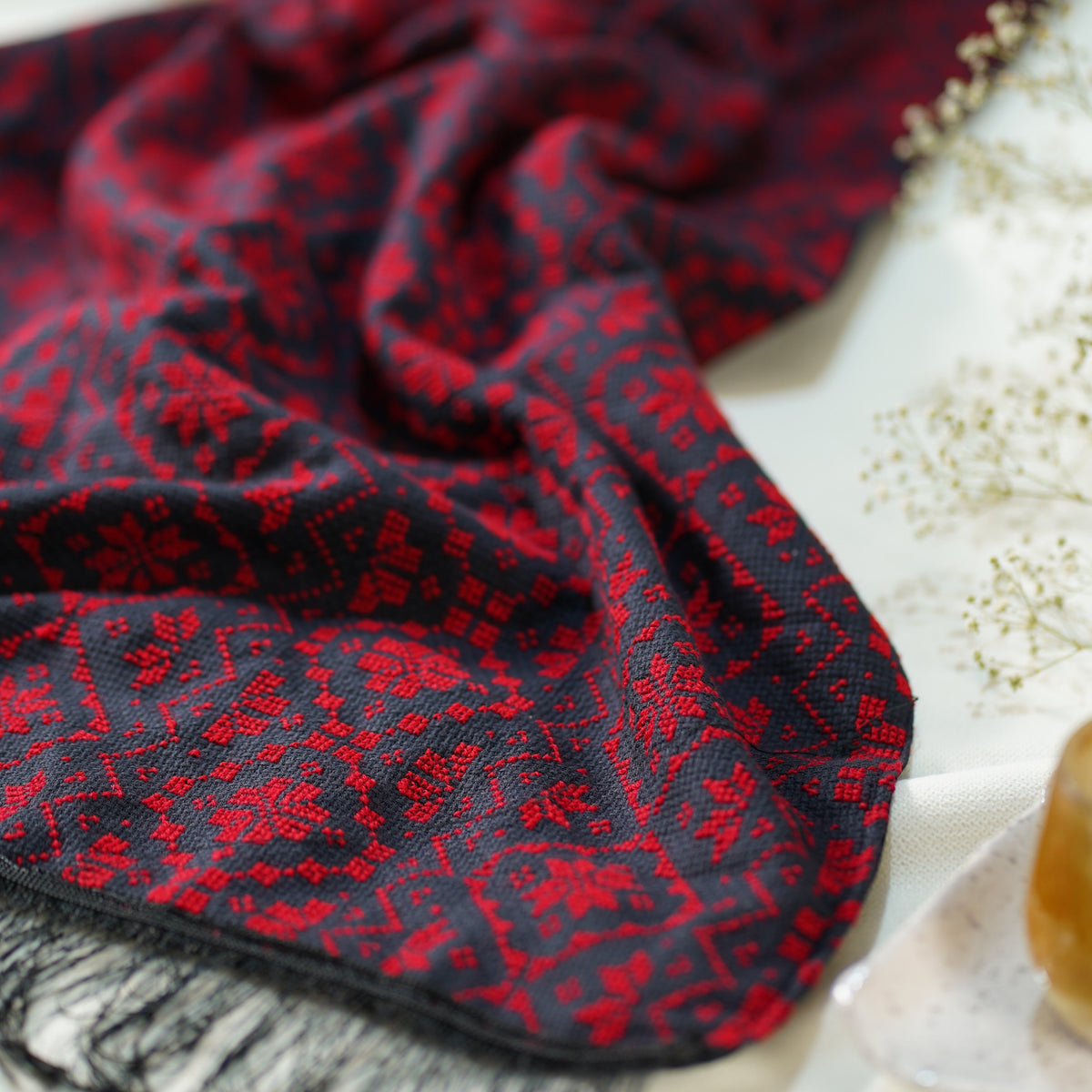 A close-up of a dark red and black hand embroidered scarf with black fringe, draped on a white surface next to small dried flowers.