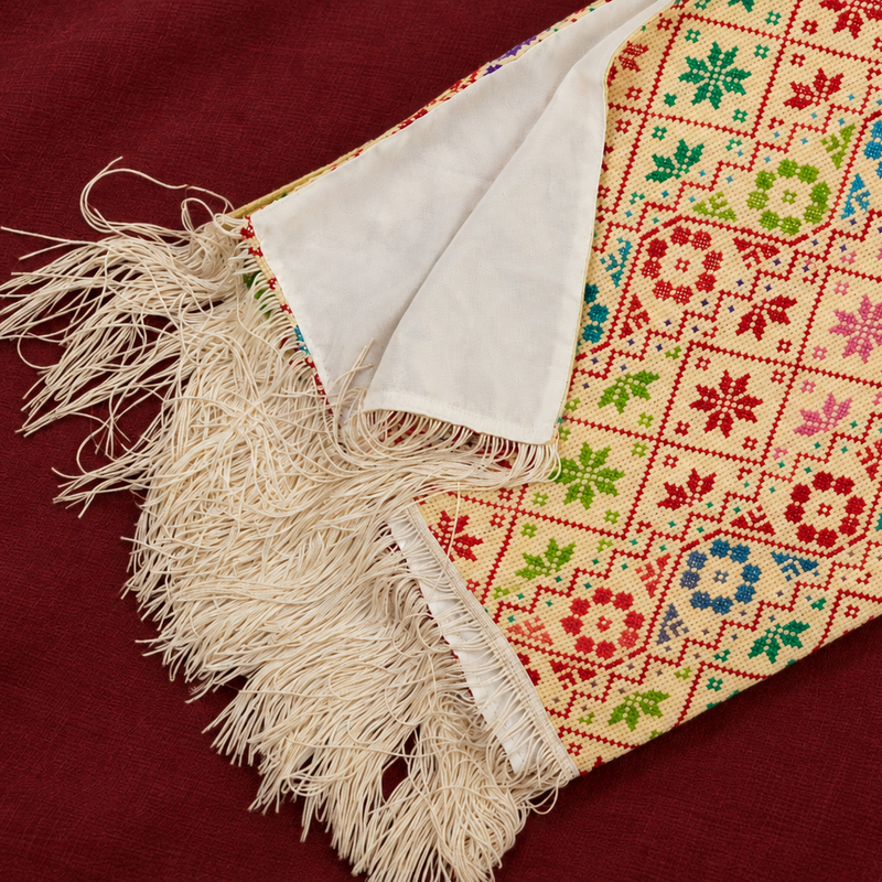 A close-up of a cream-colored fringed  shawl with a colorful embroidered geometric and floral pattern, laid diagonally on a deep red fabric background.