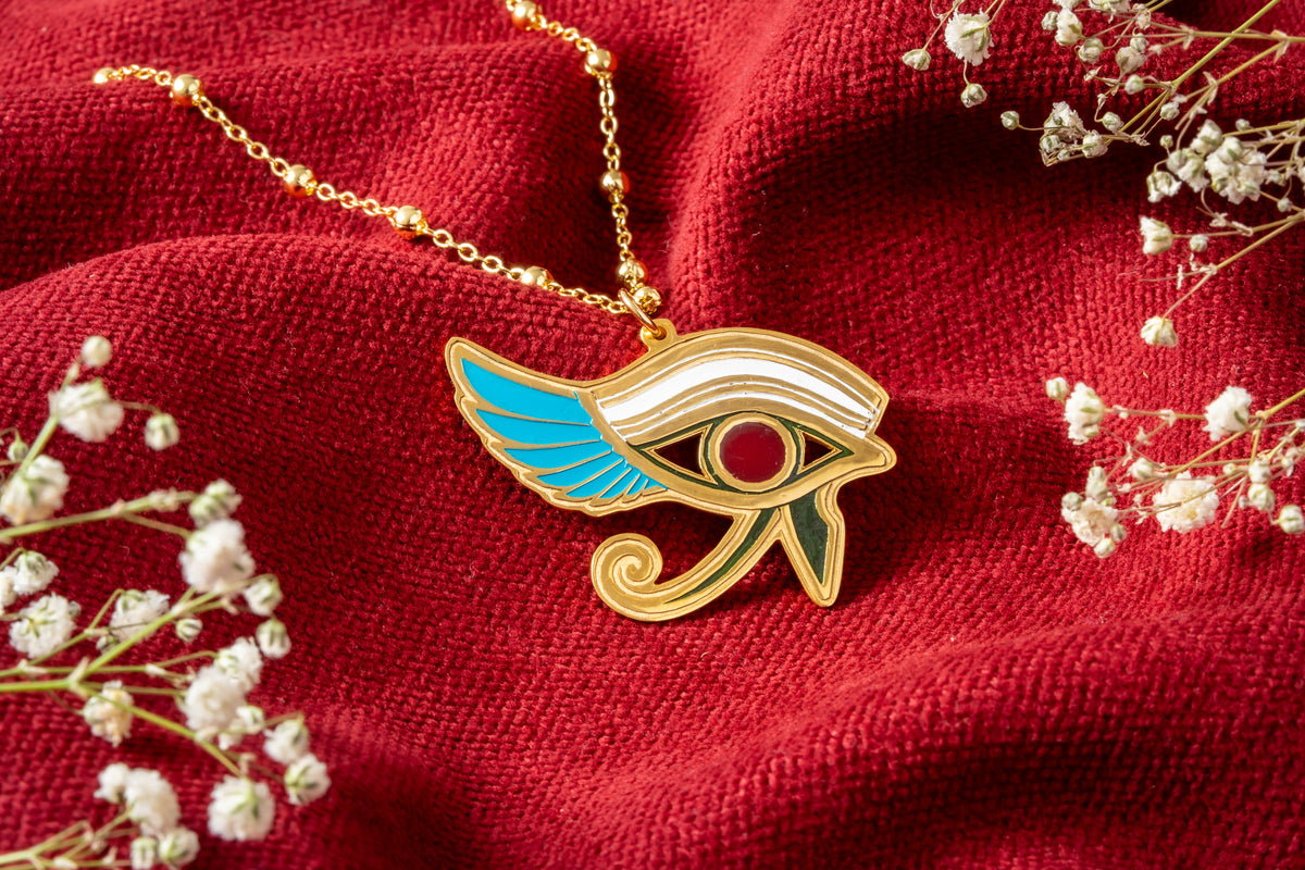 Gold-plated Egyptian Eye of Horus pendant necklace with turquoise, white, red and green enamel details, displayed on a textured red fabric background with small white baby's breath flowers.
