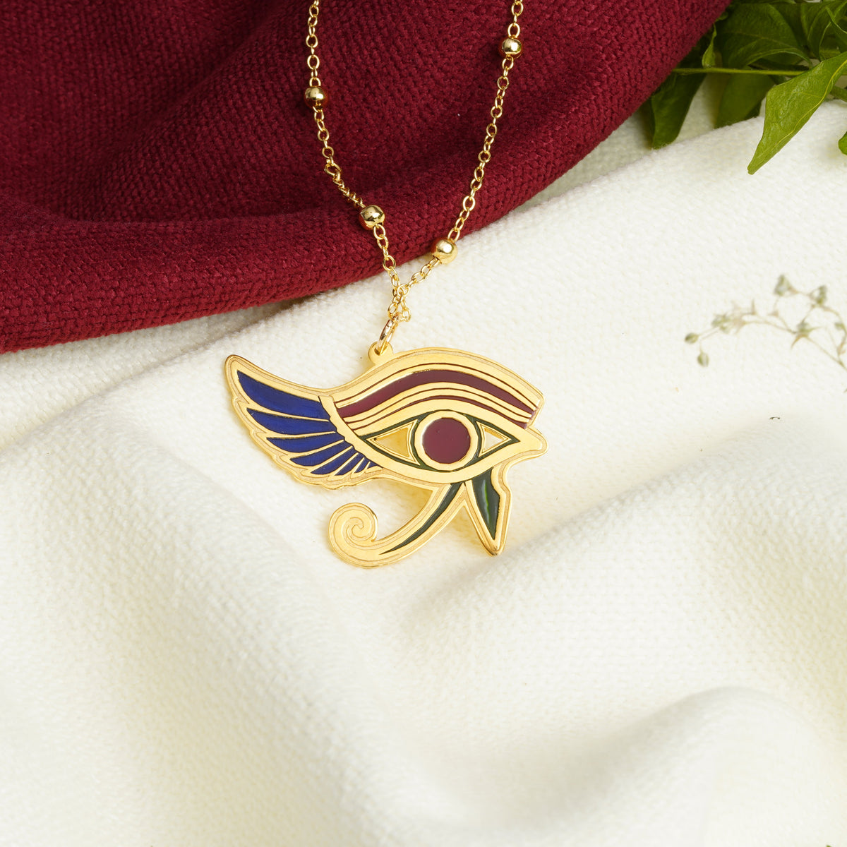 A gold pendant necklace featuring an Egyptian Eye of Horus design with blue, red, and green enamel details, displayed on a white textured fabric with a dark red cloth and green leaves in the background.