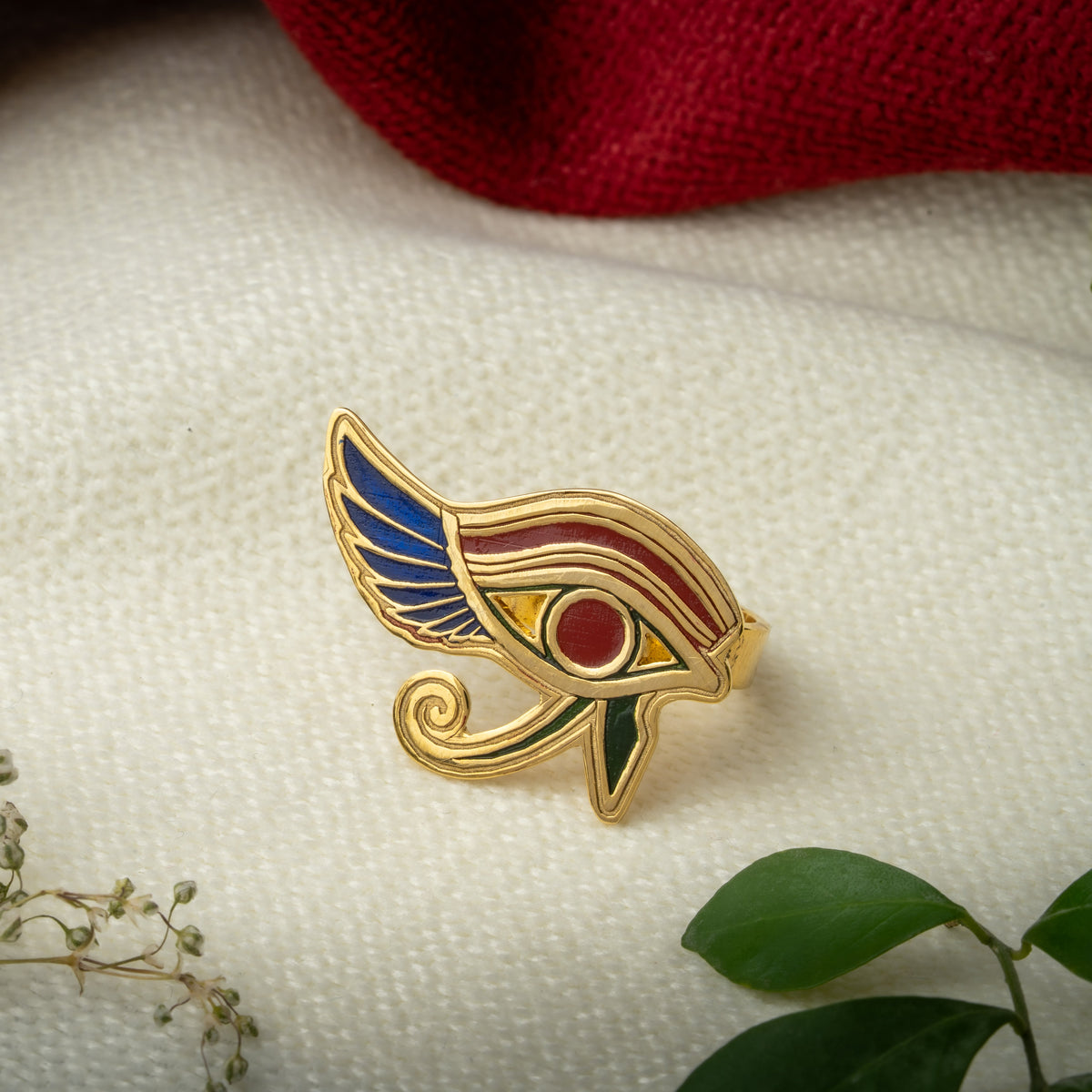 An Ancient Egyptian Eye of Horus ring with blue, red, and green enamel details, displayed on a textured white fabric with red and green decorative accents.