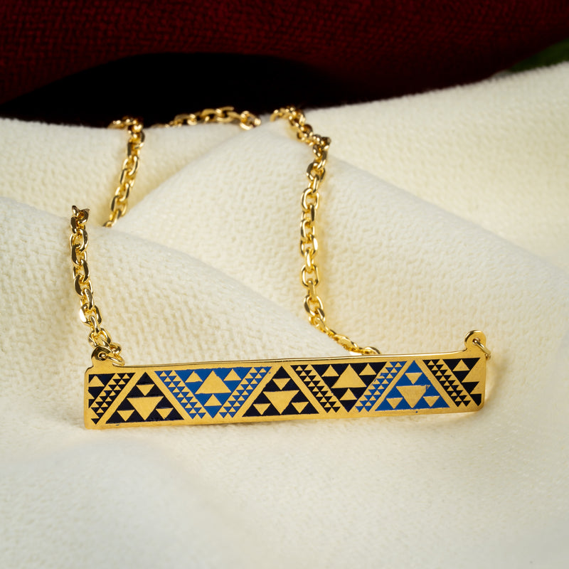 A gold bar necklace with a geometric triangle pattern in blue, navy, and gold, resting on a textured white fabric surface.