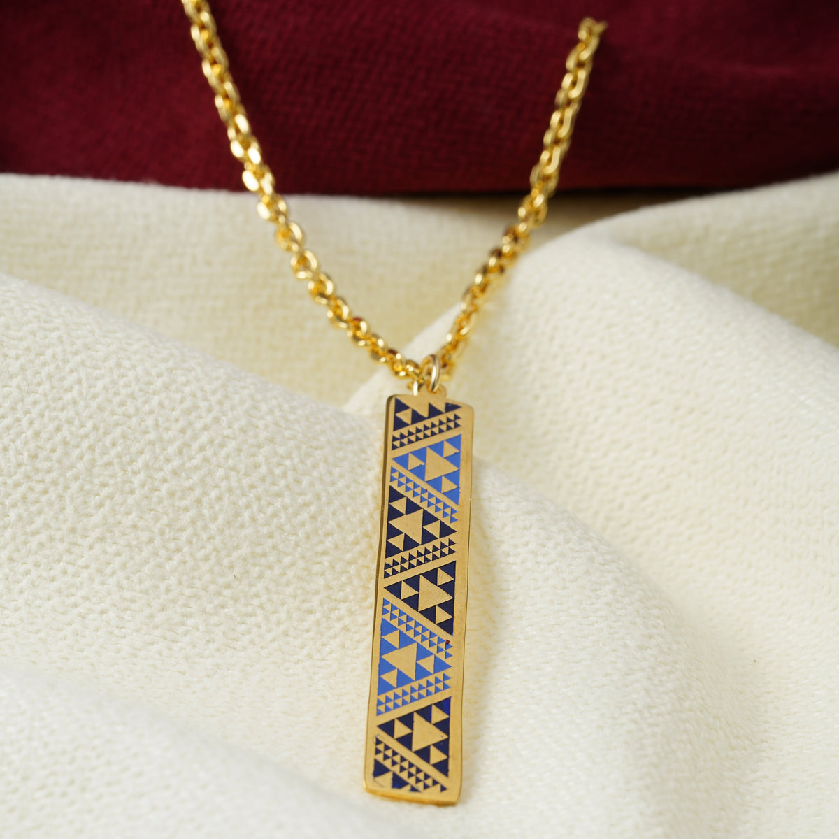 A gold chain necklace with a rectangular bar pendant featuring a blue and navy geometric pattern, displayed on draped white fabric with a dark red fabric at the top.