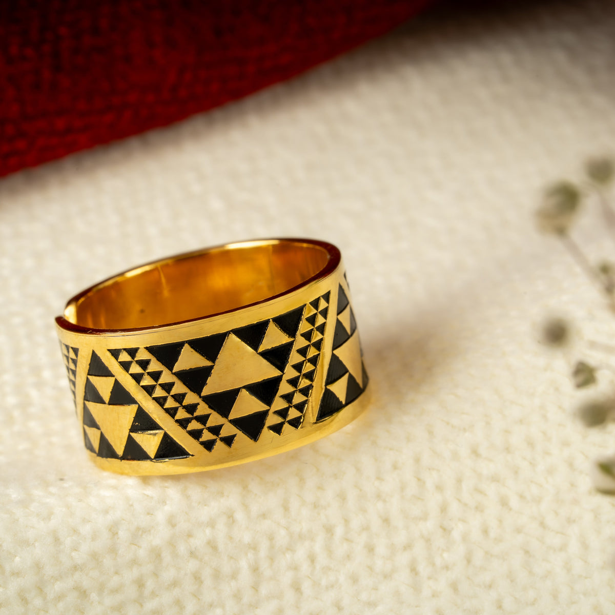 A gold band ring with a black geometric triangle pattern, resting on a textured white surface next to small dried flowers.