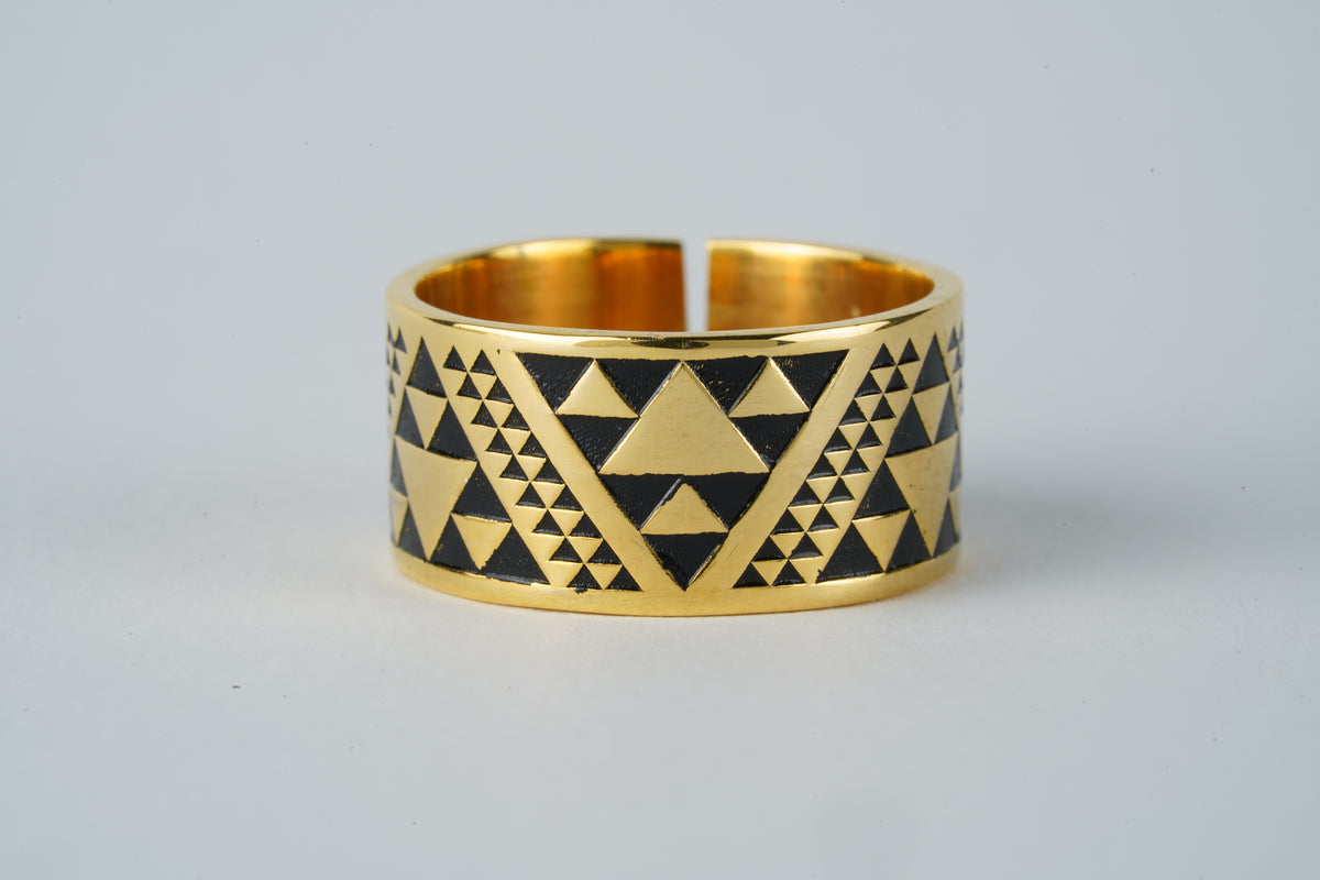 A gold plated band ring featuring a black and gold geometric triangle pattern, photographed on a neutral light gray background.