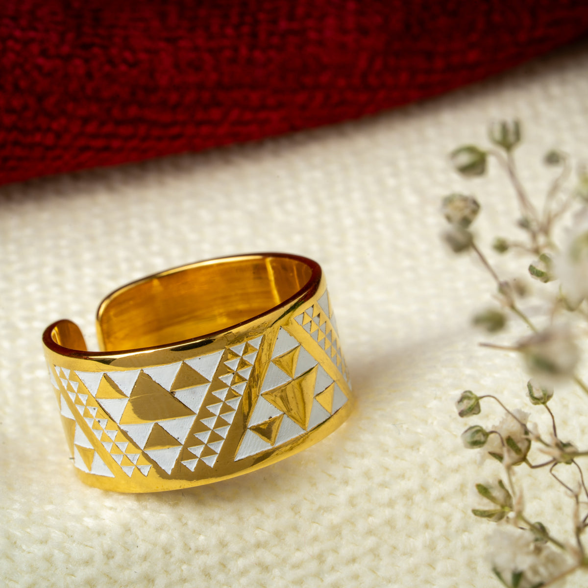 A gold-plated open band ring with white geometric triangle patterns, displayed on a textured cream fabric next to small dried flowers with a dark red background.