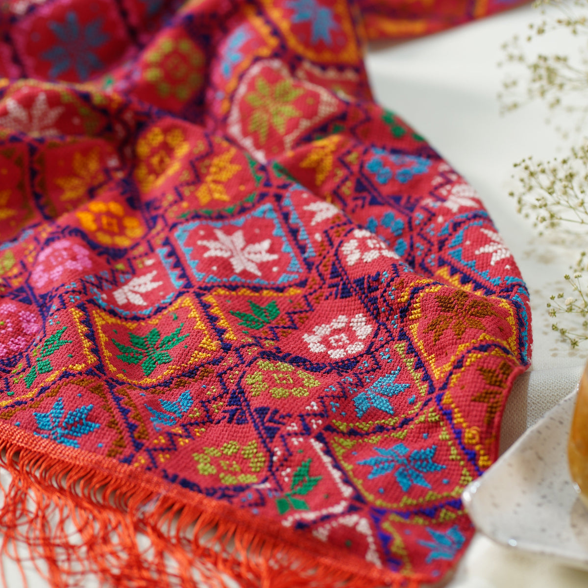 A close-up of a vibrant, multicolored hand embroidered scarf with red fringe, draped on a light surface next to dried flowers.