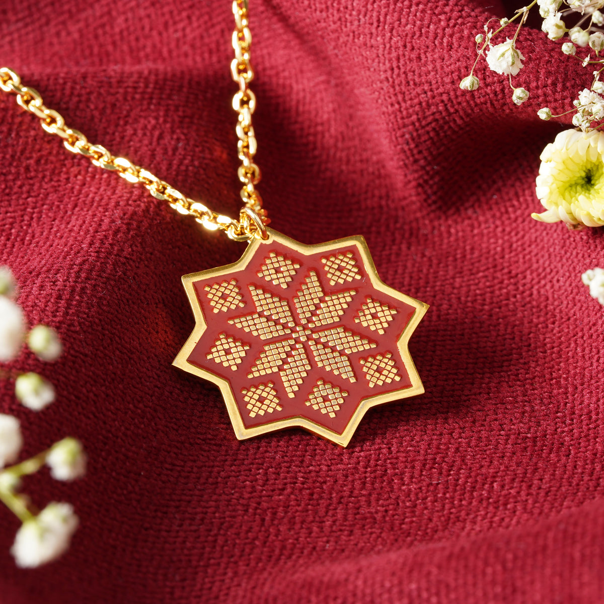A gold pendant necklace with a geometric star-shaped pendant featuring a red and gold pattern, inspired by the embroidery of Sinai Egypt, displayed on draped burgundy fabric with dried flowers and green leave