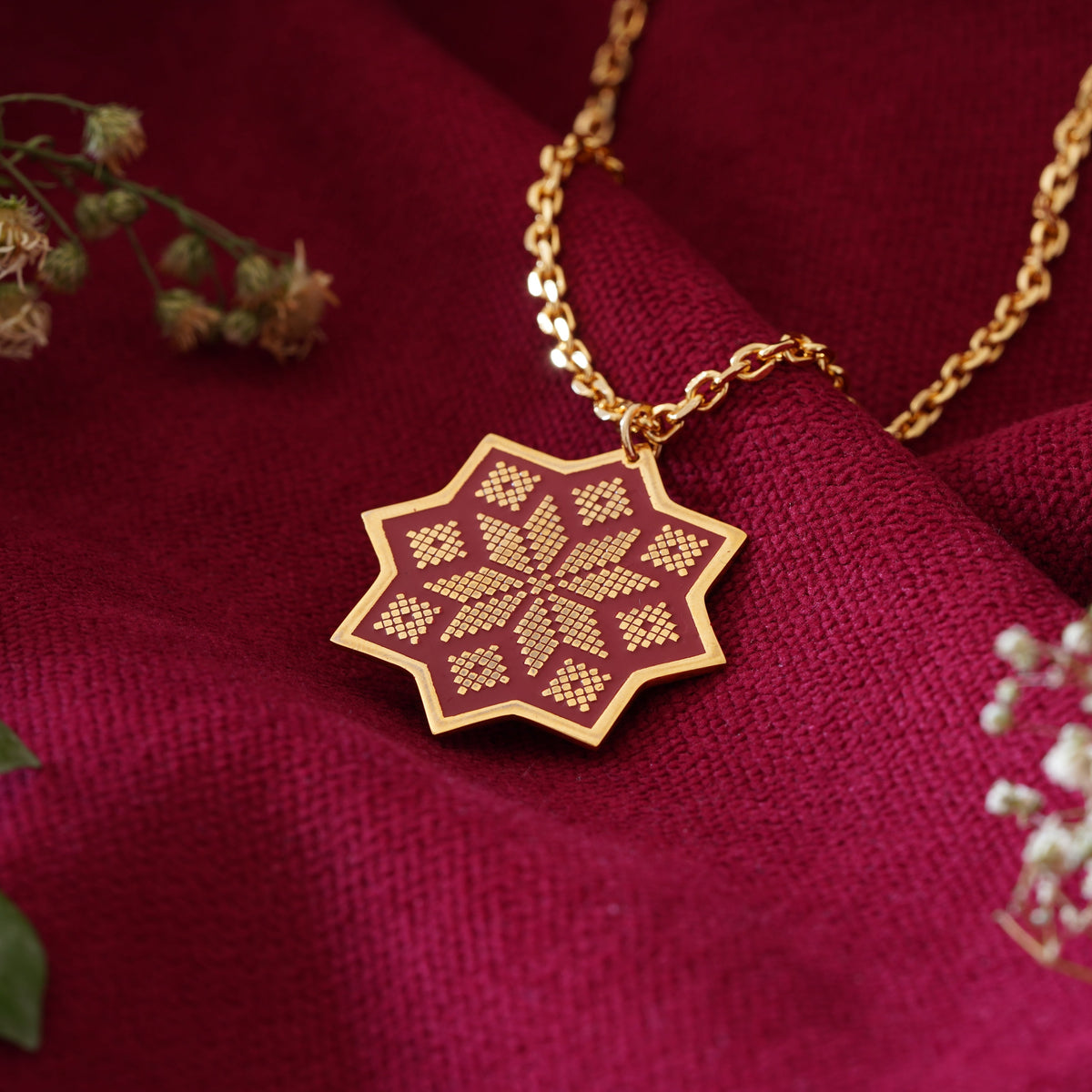 A gold pendant necklace with a geometric star-shaped pendant featuring a red and gold pattern, inspired by the embroidery of Sinai Egypt, displayed on draped burgundy fabric with dried flowers and green leave