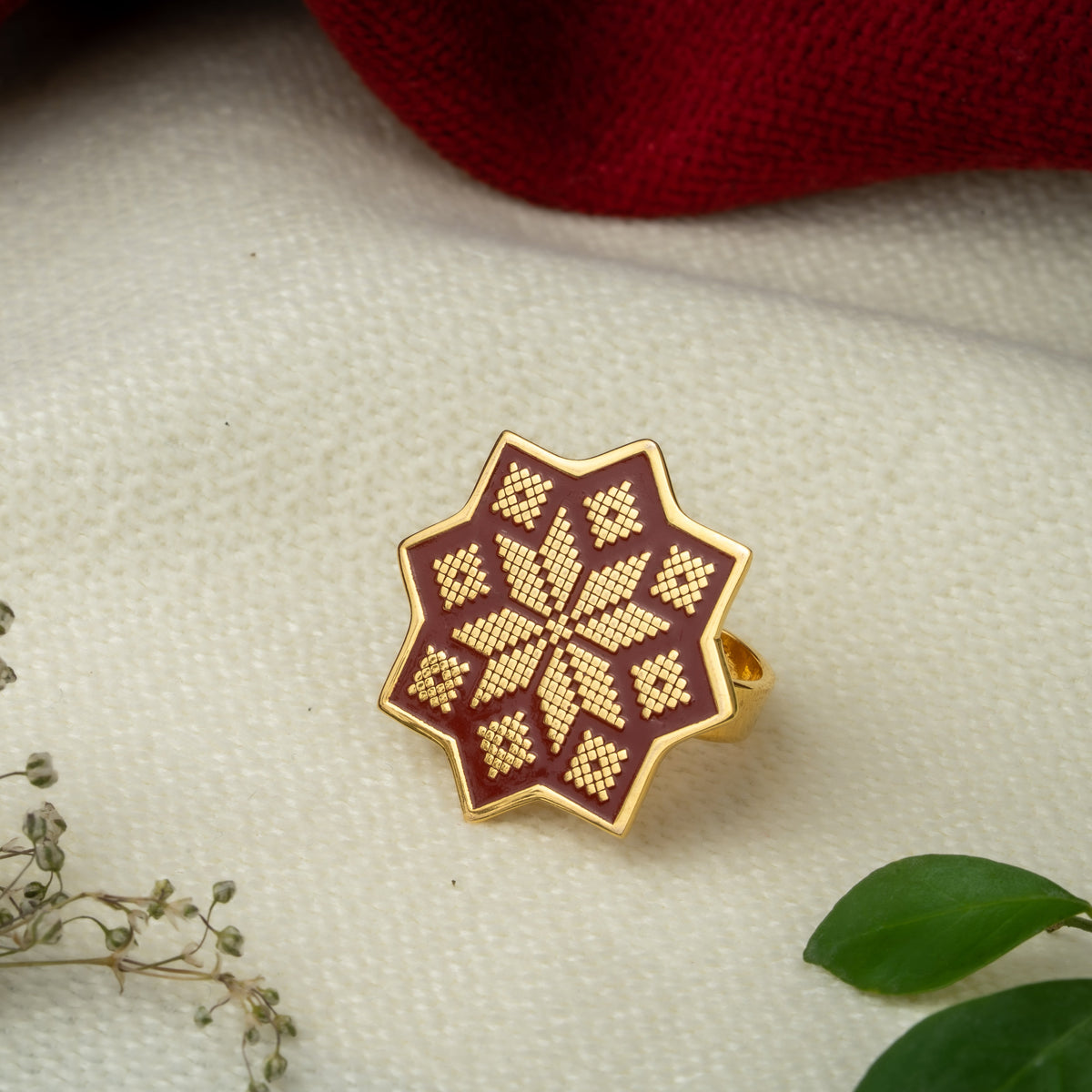 A gold ring with an eight-pointed star design featuring a red enamel details and a gold geometric cross-stitch pattern, displayed on a textured cream and maroon surface with dried flowers.