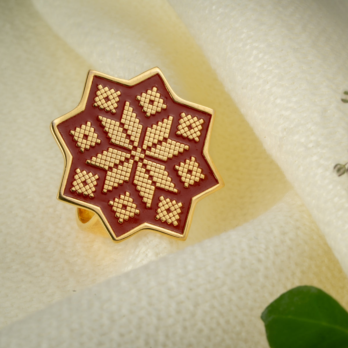 A gold ring with an eight-pointed star design featuring a red enamel details and a gold geometric cross-stitch pattern, displayed on a textured cream surface with dried flowers.