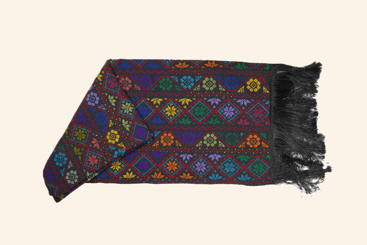 A multicolor embroidered scarf with black fringe, displayed flat against a solid cream background.