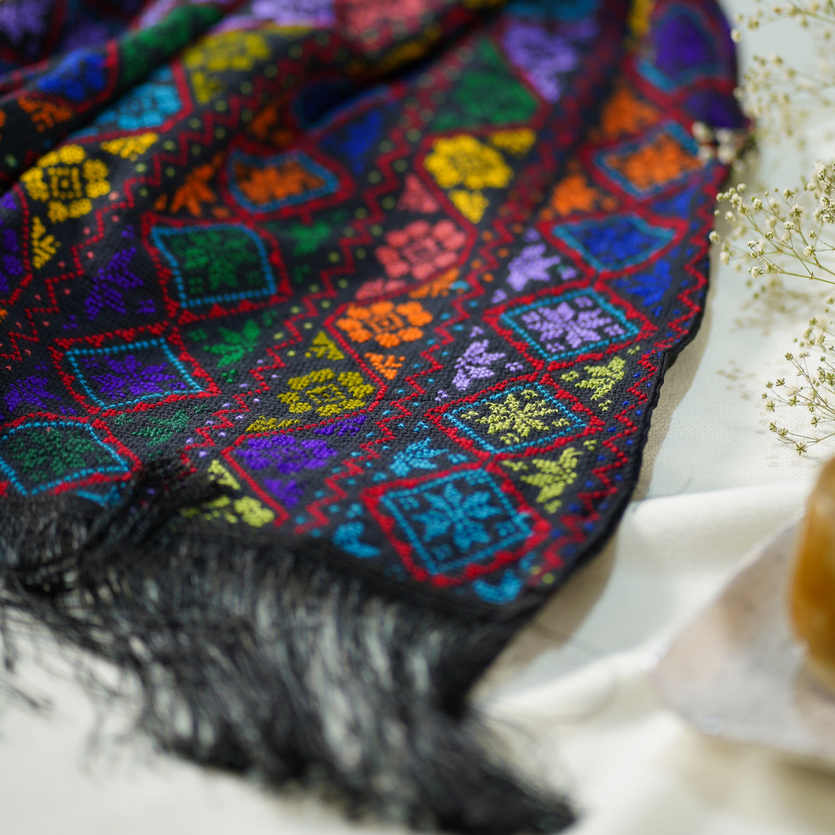 A multicolor embroidered scarf with black fringe, featuring geometric diamond patterns in red, blue, green, yellow, and purple, styled on a soft white fabric background.